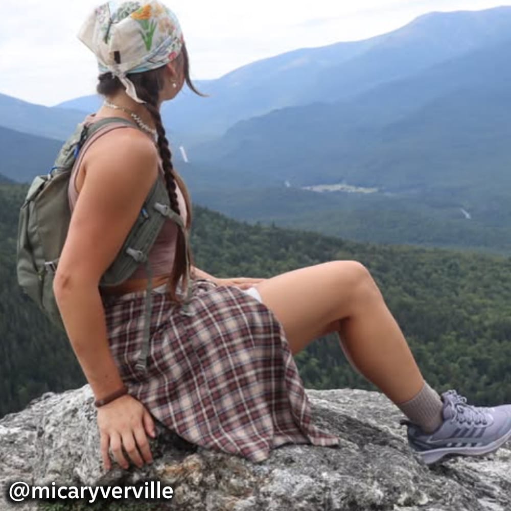 A woman sitting on a rocky overlook viewing a valley below @micaryverville. Visit Super Shoes Instagram profile.