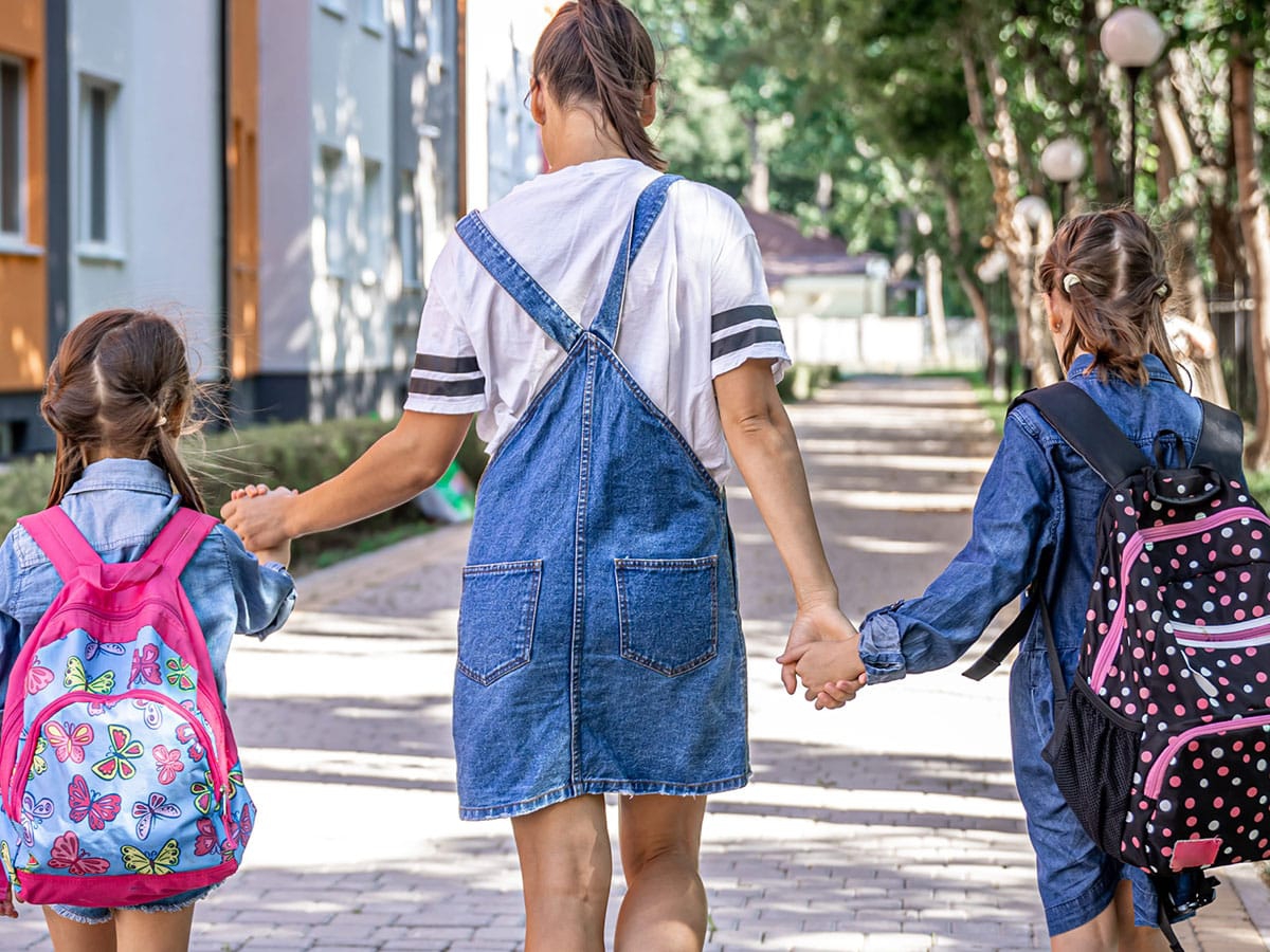 A woman walking hand in hand with two children wearing backpacks.