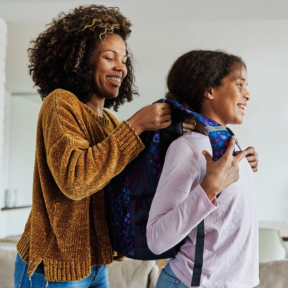 A mother helping her daughter put on a backpack.